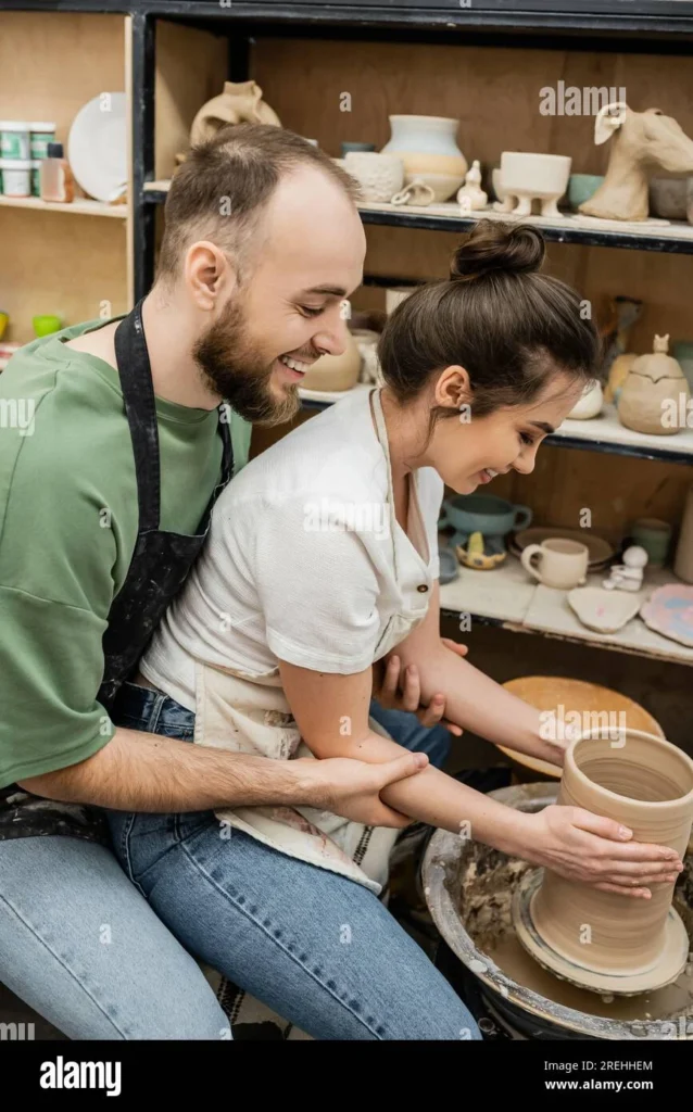 Side view of cheerful romantic artisans in aprons making clay vase on